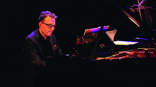 Mann in schwarzem Anzug mit Brille an einem Flügel