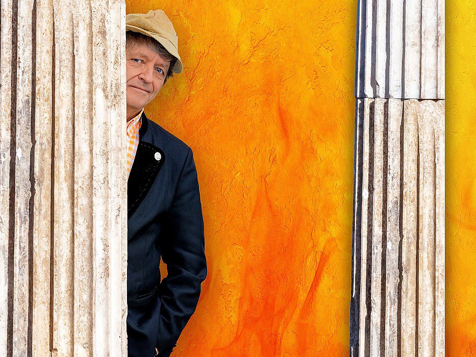 Mann mit Hut und dunklem Mantel lehnt an einer Säule vor einer orangefarbenen Wand