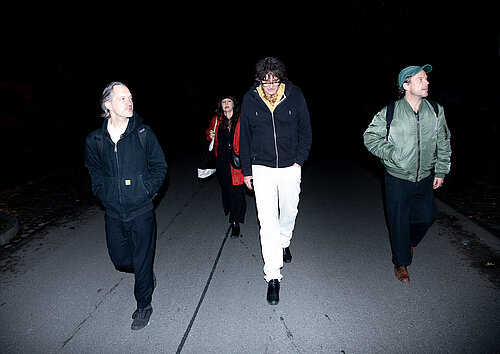 Vier Personen gehen auf einer nächtlichen Straße, zwei tragen helle Hosen und Jacken, eine Person hat einen Rucksack.