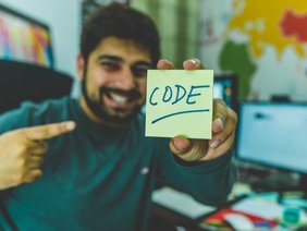 Junger Mann hält lächelnd einen Notizzettel in die Kamera, auf dem das Wort "Code" geschrieben steht