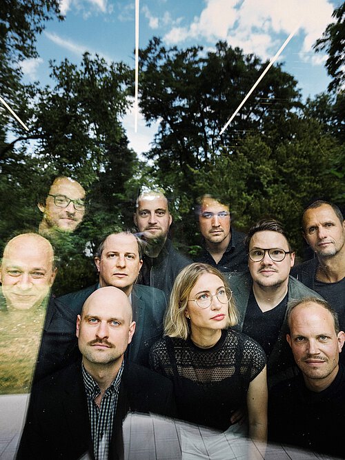 Gruppe von Menschen im Freien vor Bäumen und blauem Himmel, einige tragen dunkle Kleidung.