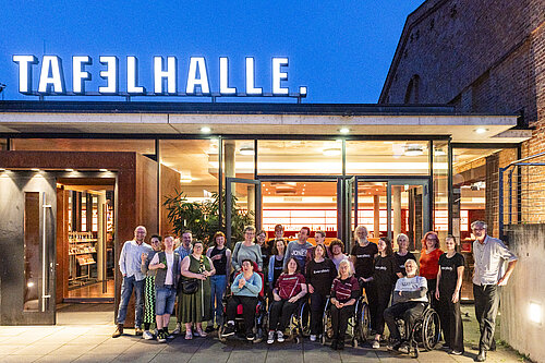 Gruppe von Menschen, darunter mehrere im Rollstuhl, steht vor einem Gebäude mit dem beleuchteten Schriftzug 'tafelhalle'