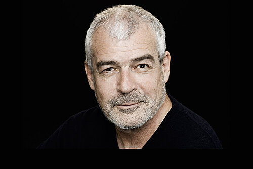 Älterer Mann mit grauen Haaren und Bart, trägt schwarzen Pullover, vor schwarzem Hintergrund.