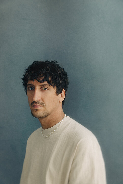 ein Mann mit weißem Shirt vor blauem Hintergrund, guckt in die Kamera