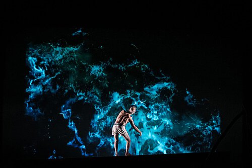 Ein Mann oberkörperfrei steht mit dem Rücken zum Publikum, im Hintergrund ist eine Wasseranimation projiziert.