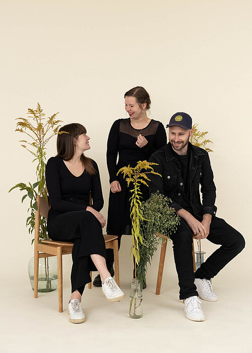 drei junge Menschen in schwarz gekleidet vor weißem Hintergrund, zwei davon sitzen
