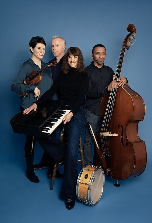 Vor einem blauen Hintergrund stehen vier Musiker*innen: Eine Frau mit Violine im Arm, eine Frau mit einem kleinen Flügel, ein Mann mit einem Kontrabass und ein weitere Mann. 