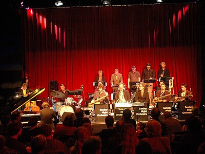 Jazzorchester auf Bühne mit roten Vorhängen, Musiker spielen verschiedene Instrumente wie Klavier, Schlagzeug und Blasinstrumente.