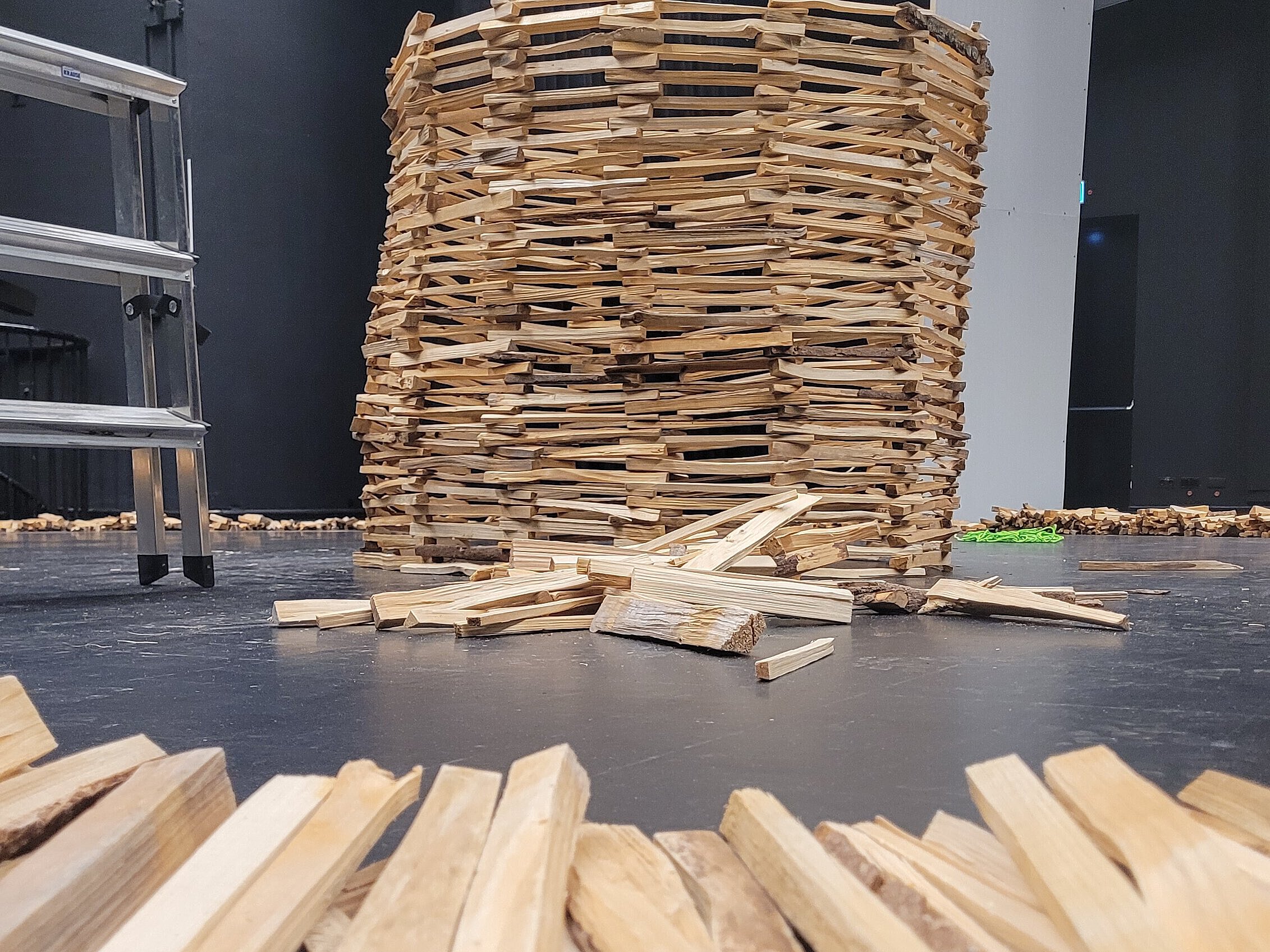 Turm aus übereinander gestapelten Holzklötzen auf schwarzem Boden, im Vordergrund verstreute Holzstücke, im Hintergrund Leiter und schwarze Vorhänge.