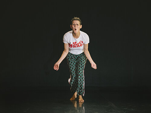 Person steht barfuß, trägt weißes T-Shirt mit rotem Schriftzug 'Let's Dance' und gemusterte Hose, vor schwarzem Hintergrund.