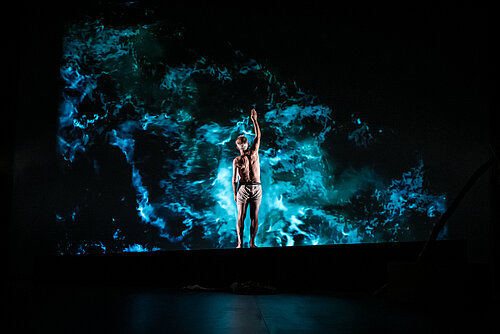 ein Mann in Boxershorts steht vor einem, an die Wand projiziertem Abbild des Meeres