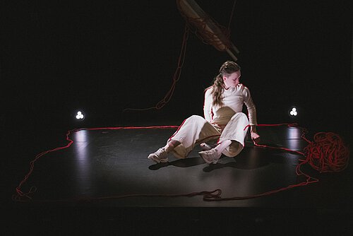 Eine Frau sitzt auf schwarzem Bühnenboden, umgeben von einem roten Seil, rechts neben ihr ein großes rote Wollknäuel