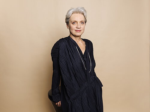 Person mit kurzen weißen Haaren trägt ein langärmeliges, schwarzes Kleid und lehnt an einer beigen Wand.