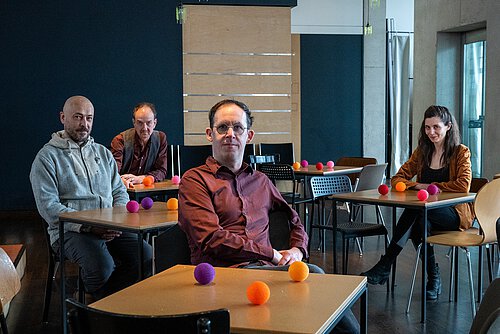 Drei Männer und eine Frau sitzen an verschiedenen kleinen Tischen. Auf jedem Tisch sitzen kleine, bunte Bälle. 