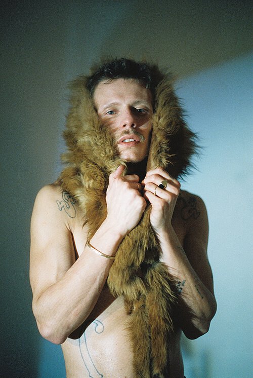 ein Mann mit nacktem Oberkörper blickt in die Kamera. Er hält ein Fell über den Kopf