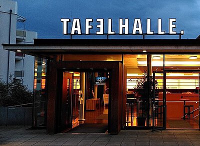 Beleuchteter Eingang der Tafelhalle am Abend mit Glasfront und offenem Türbereich.