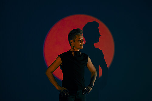 Person mit hochgestecktem Haar und schwarzem ärmellosem Oberteil vor rotem Kreis auf dunklem Hintergrund, Hände in Hüftposition, Schatten an der Wand sichtbar.