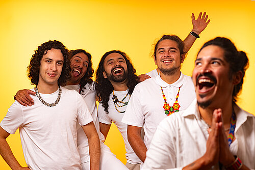 Gruppenfoto der Band vor einem gelben Hintergrund, sie tragen weiße Poloshirts und manche von ihnen bunte Ketten, sie lachen