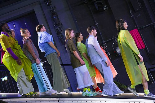 Person in rotem Oberteil sitzt auf einem weißen Tuch, das von mehreren Personen gehalten wird, auf einer weißen Bühne in dunklem Raum