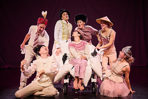 Sieben Personen in bunten Kostümen posieren auf einer Bühne, eine Person sitzt in einem Rollstuhl, die anderen stehen oder knien um sie herum.