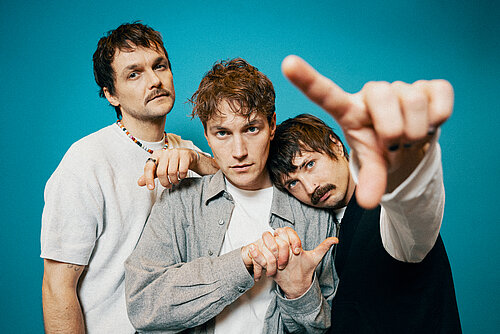 Drei Männer in lässiger Kleidung stehen vor einem türkisem Hintergrund