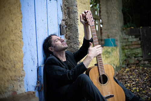 Person in schwarzer Kleidung sitzt an eine blaue Holztür gelehnt und spielt auf einer akustischen Gitarre aus Holz.