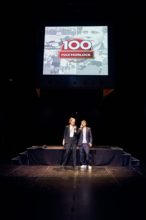 zwei Personen auf der Bühne singen, über ihnen eine Leinwand
