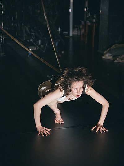 Person mit lockigem Haar in weißem Oberteil und beiger Hose in kauernder Haltung auf dunklem Boden, an Seilen befestigt