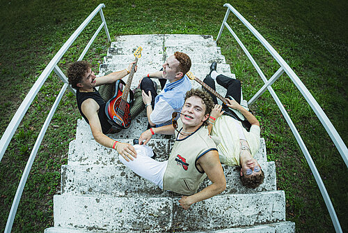 Vier Personen sitzen auf einer steinernen Treppe im Freien, zwei davon halten E-Gitarren