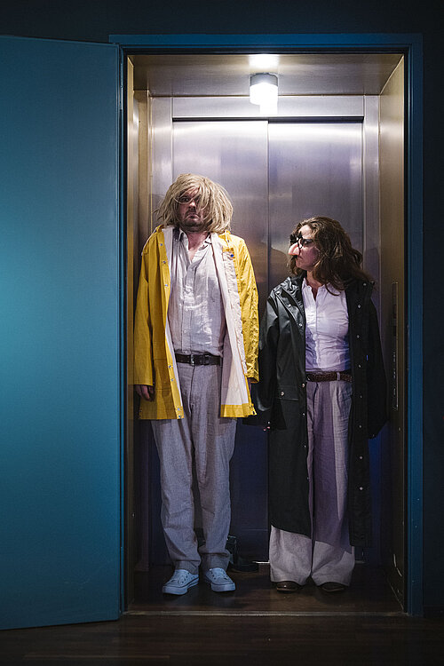 Zwei Personen stehen in einem Aufzug mit Metallwänden, eine trägt einen gelben Mantel, die andere einen dunklen Mantel.