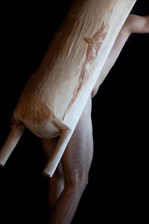 Teilweise sichtbarer nackter Mann hält ein großes, längliches, helles Holzobjekt mit zwei kurzen Beinen vor einem schwarzen Hintergrund.