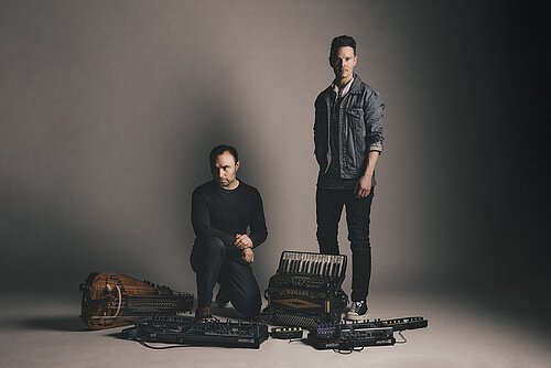 Zwei Männer mit Musikinstrumenten und elektronischem Equipment vor neutralem Hintergrund, einer steht, der andere kniet.