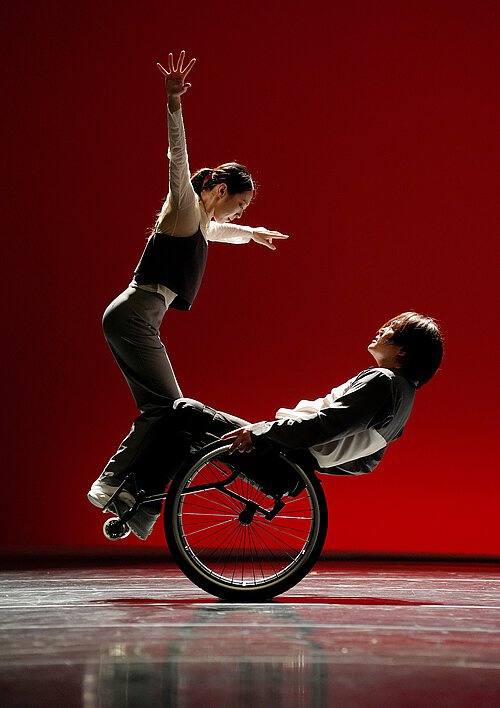 Zwei Tänzer auf einer Bühne vor rotem Hintergrund, einer sitzt im Rollstuhl, der andere steht auf dessen Oberschenkeln mit erhobenen Armen