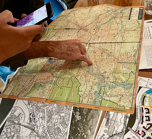 Hand zeigt auf eine aufgeklappte Landkarte, daneben Smartphone mit Karten-App, darunter weitere Karten und Stadtpläne auf einem Holztisch