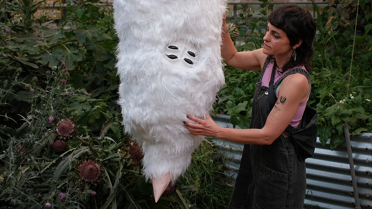 Person in schwarzem Latzkleid berührt eine große, hängende, weiße Vogelskulptur in einem Gewächshaus