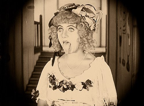 eine Frau mit gelocktem Haar blickt an der Kamera vorbei und streckt dabei ihre Zunge raus