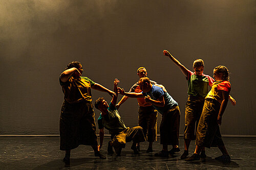 Sechs Personen in bunter, lockerer Kleidung tanzen auf einer Bühne mit dunklem Hintergrund und warmem Licht