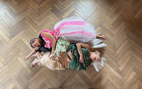 Drei Frauen liegen eng aneinander geschmiegt auf einem Parkettboden, tragen bunte, fließende Kleider in Rosa, Beige und Grün.