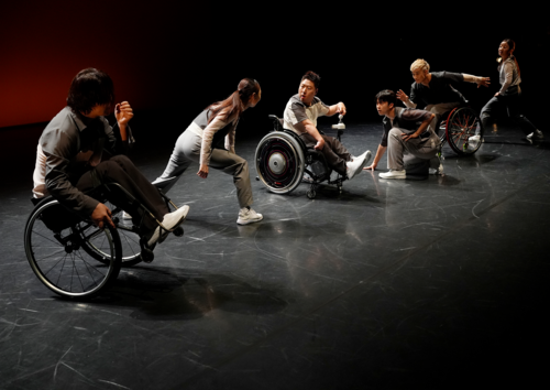 Sechs Personen, darunter zwei im Rollstuhl, führen auf einer Bühne eine Tanzperformance mit dynamischen Bewegungen aus