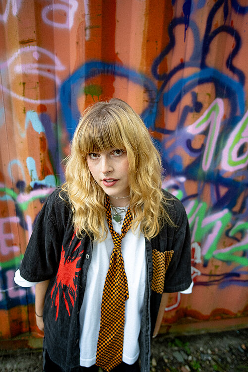 eine junge Frau mit offenem Hemd vor einem bunt angesprühten Container