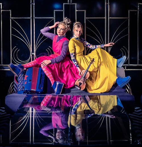 zwei Frauen in pinkem und gelben Kleid mit Violine und Akkordeon Rücken an Rücken sitzend