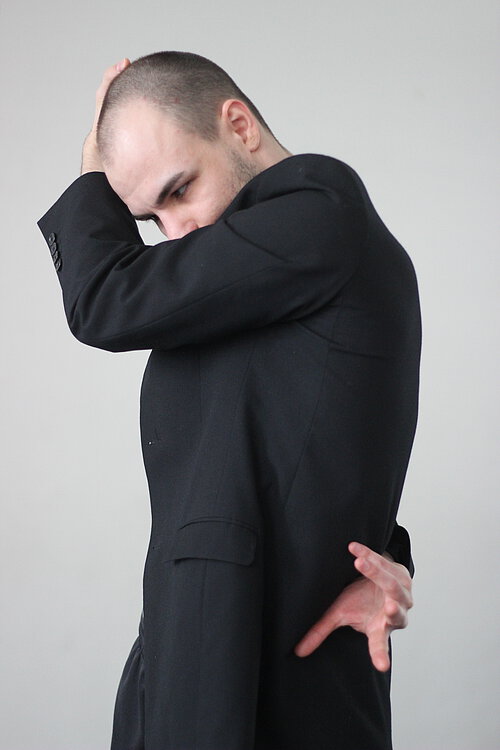 Mann mit schwarzem Anzug und schütterem Haar, hät den linken Arm vors Gesicht