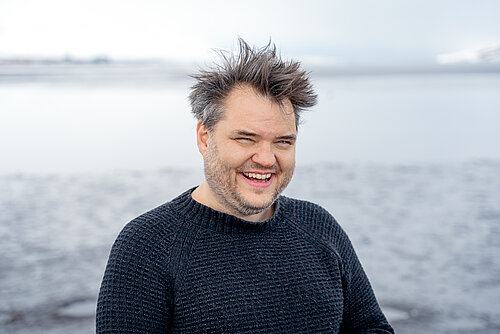 Mann mit verwuscheltem Haar lachend in dunklem Pullover