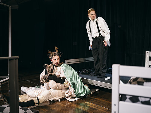 Mann in Käferkostüm sitzend vor metallenem Bettgestell, auf dem Bettgestell eine Frau in Anzug 