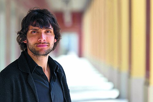 Mann mit dunklem Hemd und schwarzer Jacke steht in einem Gang mit gelben Säulen und weißem Boden.