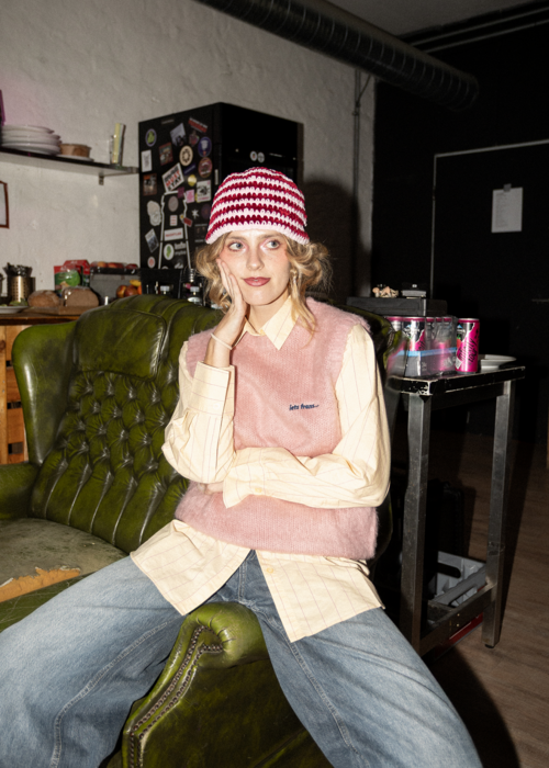 Person sitzt mit gespreizten Beinen auf einem grünen Ledersessel und trägt eine rot-weiß gemusterte Mütze, eine rosa Strickweste über einem hellen Hemd und blaue Jeans