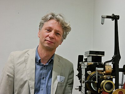 Ein Mann mit hellgrauem Sakko und blauem Hemd steht neben einer alten Filmprojektionsmaschine mit sichtbaren Zahnrädern und Hebeln