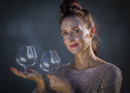 Frau in glitzerndem Kleid und mit Glitzer im Gesicht mit zwei Weingläsern in den Händen