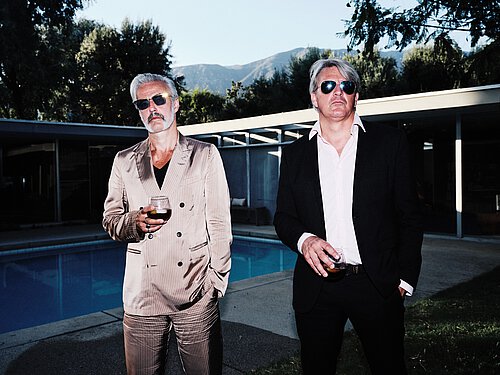 Zwei Männer in Anzügen stehen nebeneinander an einem Pool, jeder hält ein Glas in der Hand, im Hintergrund Berge und ein modernes Gebäude