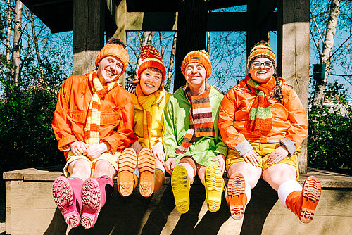 Vier Personen in bunten Regenjacken, Mützen und Schals sitzen nebeneinander auf einer Holzbank und strecken ihre Gummistiefel nach vorne